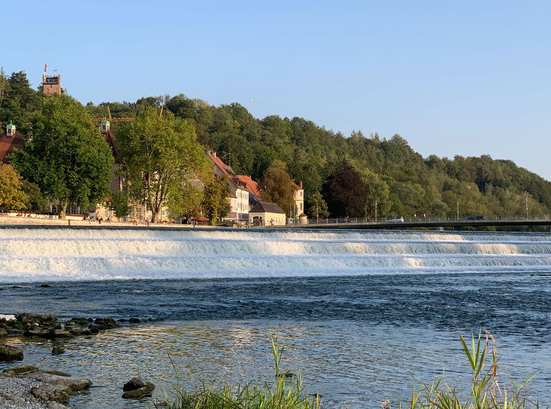 Lechwehr Landsberg und Flussufer im Vordergrund. Lechmauer Häuser und Bäume im Hintergrund. Blauer Sommerhimmel